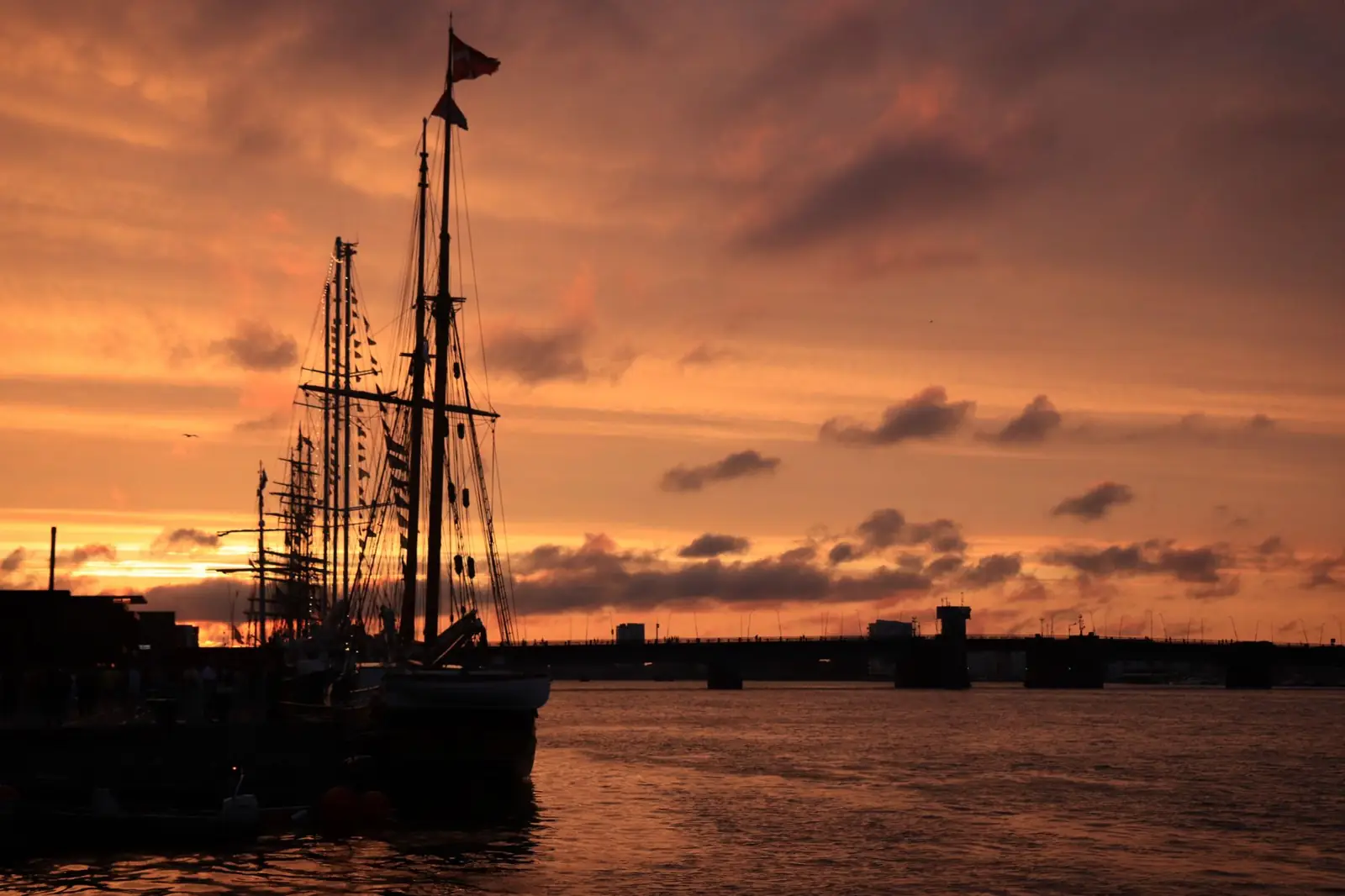 A serene sunset over Aalborg harbor with silhouetted sailing ships against a vibrant sky.