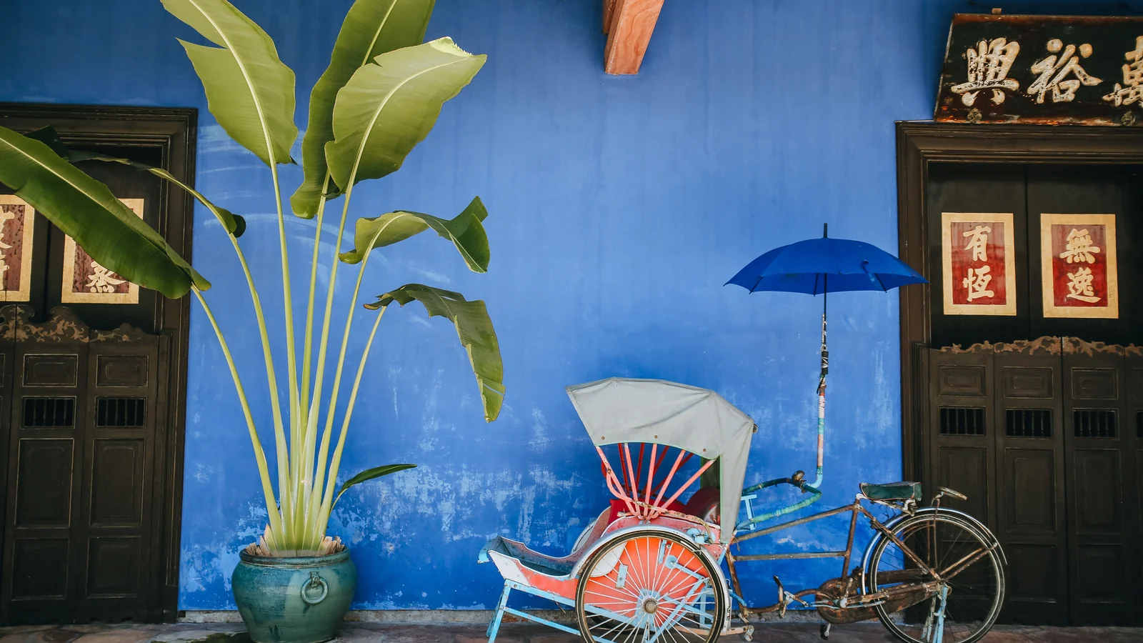 A colourful trishaw and potted plant against the Blue Mansion’s indigo wall and wooden doors.
