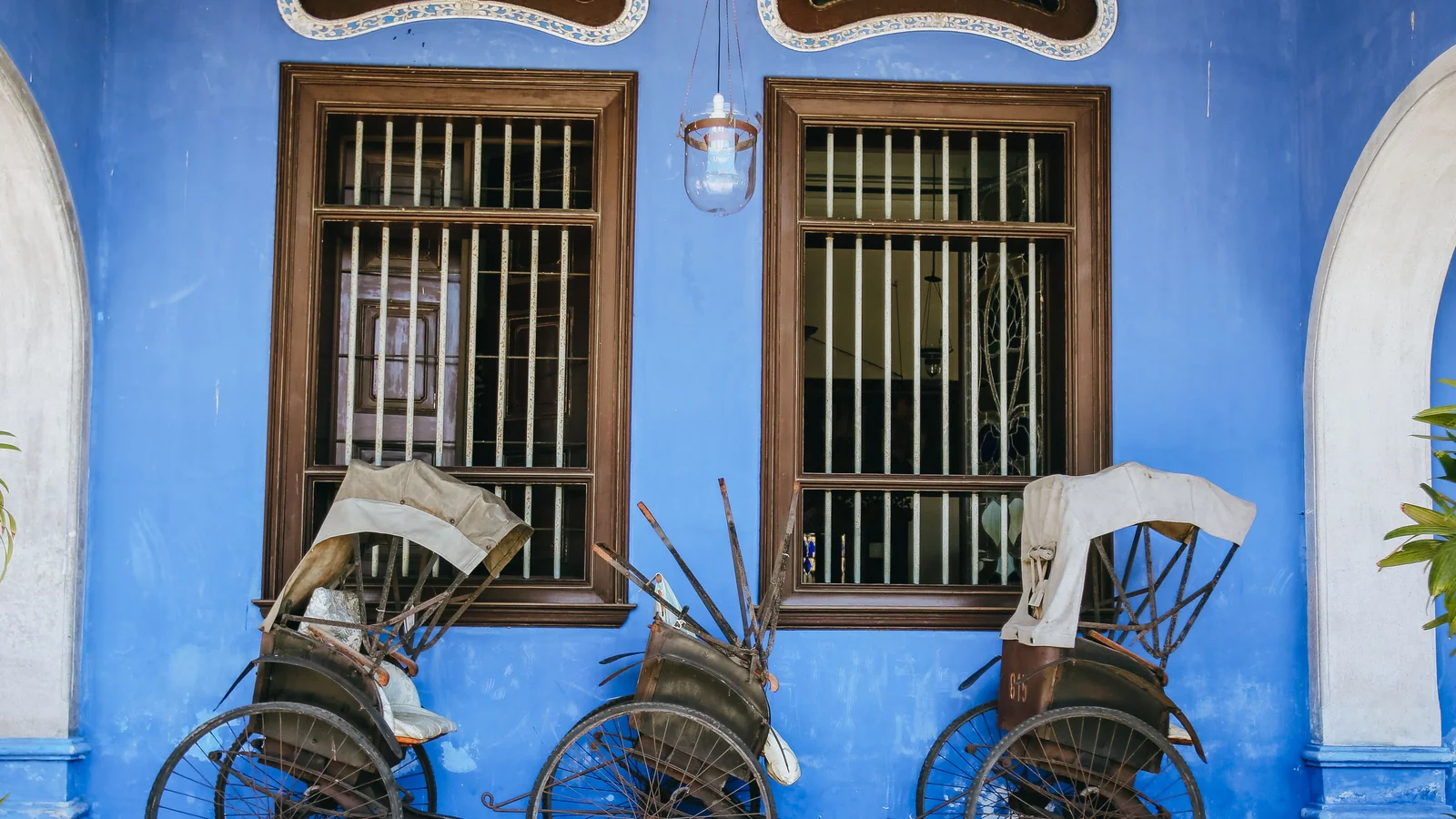 Two traditional trishaws beneath wooden windows against the Blue Mansion’s indigo walls in George Town.