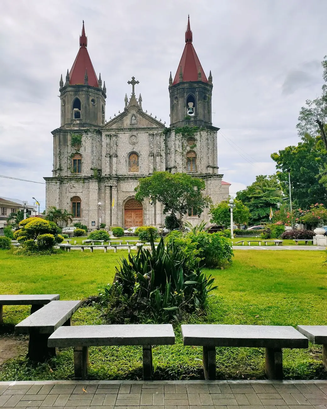 Stone façade and twin red spires of Molo Church seen across the planted plaza in Iloilo City