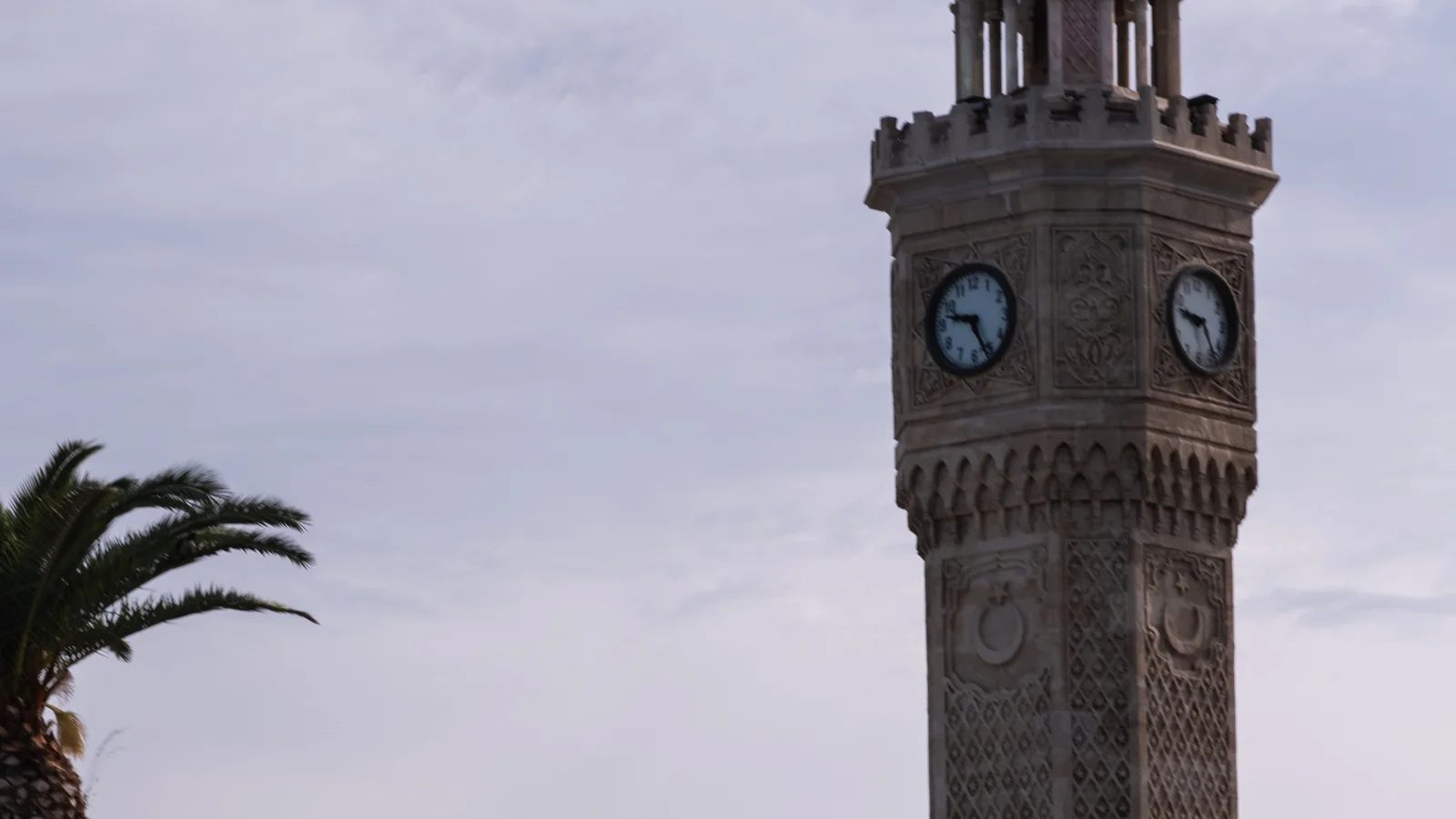 Clock faces and carved stonework on İzmir’s Clock Tower beside a palm in Konak Square.
