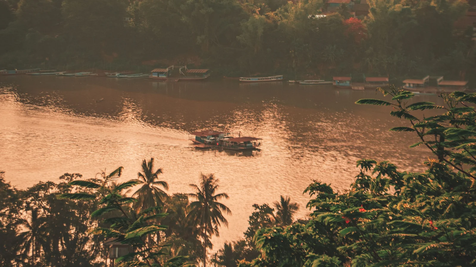 A boat on the sunlit Mekong in Luang Prabang, with palms and houses on the banks.