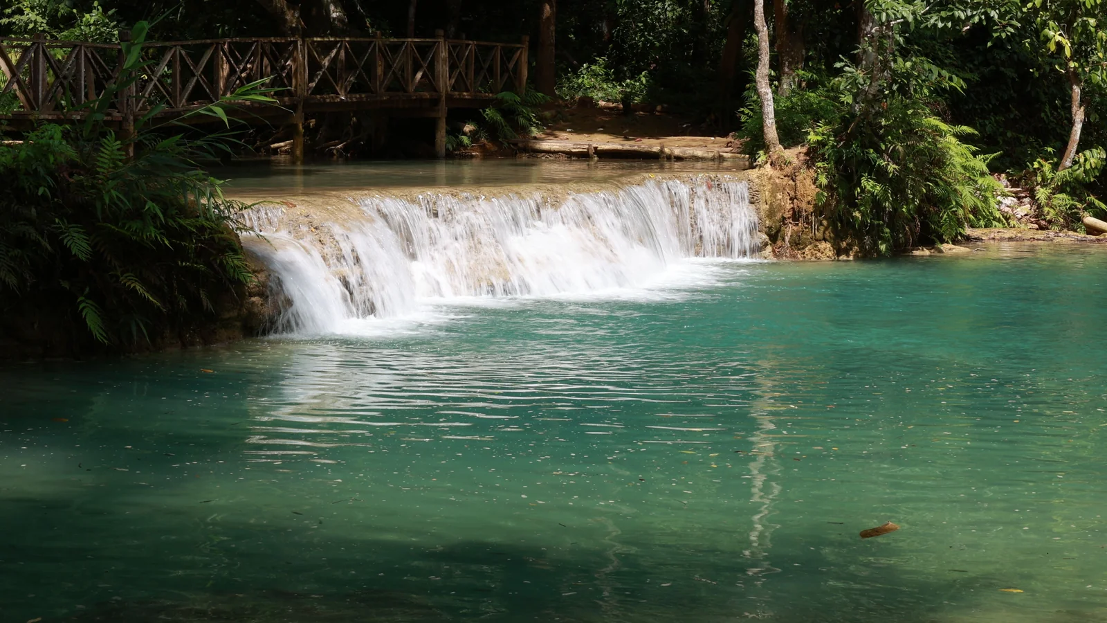 A limestone cascade and green-blue pool below a wooden bridge at Kuang Si Falls.