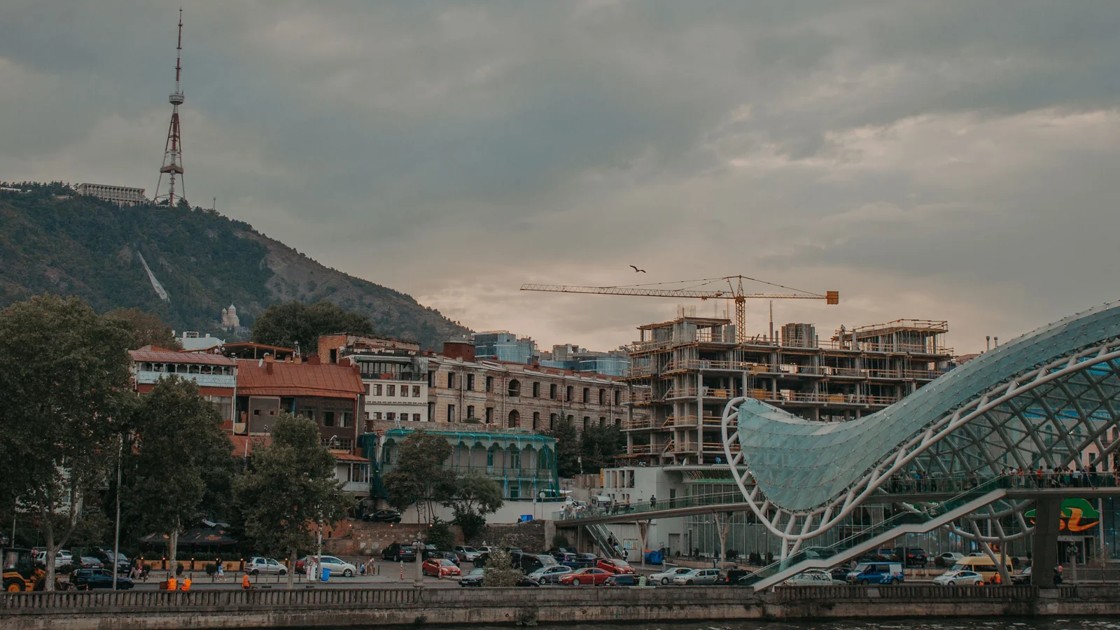 The glass canopy of the Bridge of Peace beside the Mtkvari River, with Tbilisi’s hillside television tower beyond.