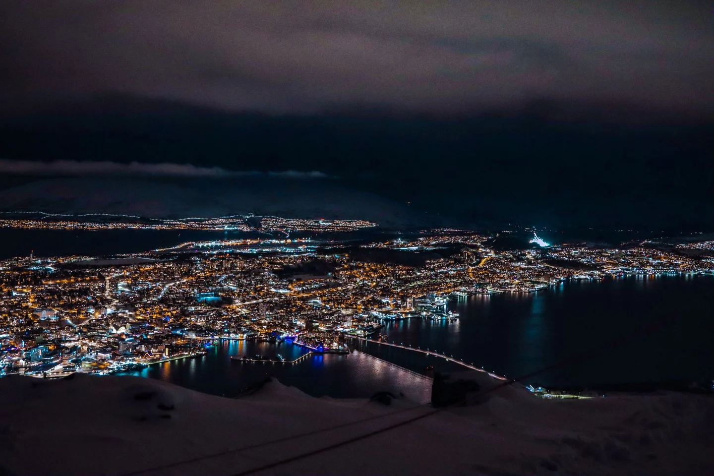 Tromsø’s illuminated harbour and bridge seen from the snowy hillside at night