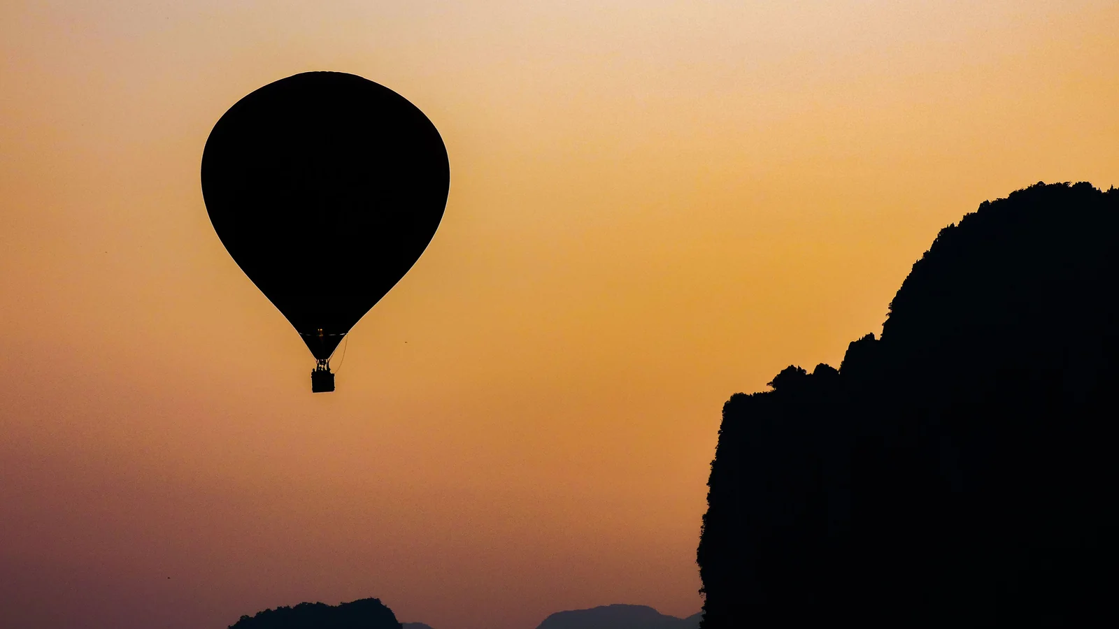 A hot-air balloon silhouetted against an orange sky beside Vang Vieng’s limestone cliffs.