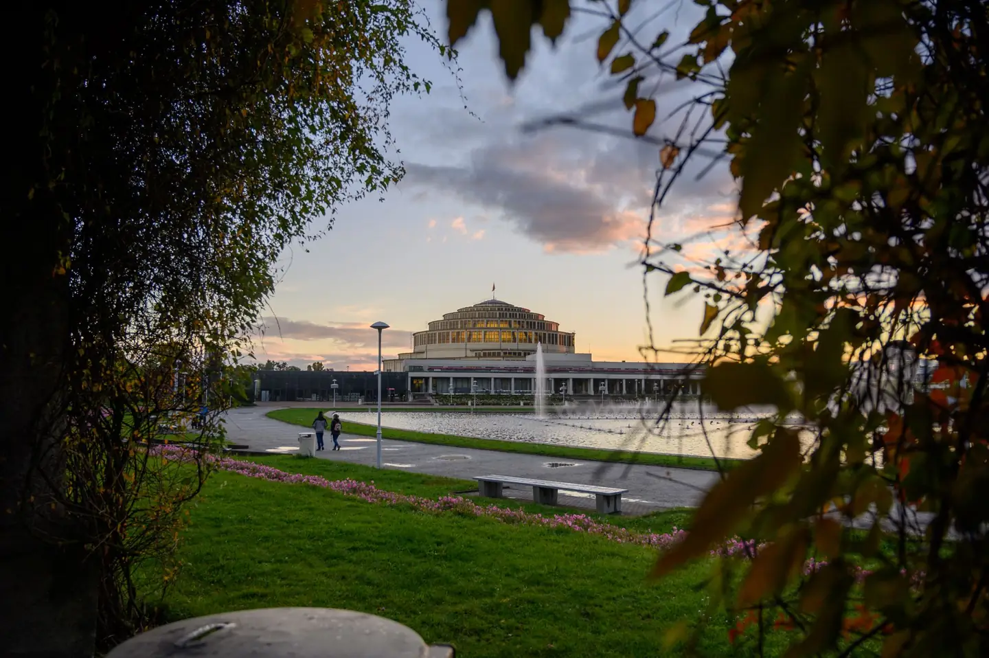 Wrocław’s Centennial Hall dome beyond the Pergola pond and fountain basin, framed by trees at sunset