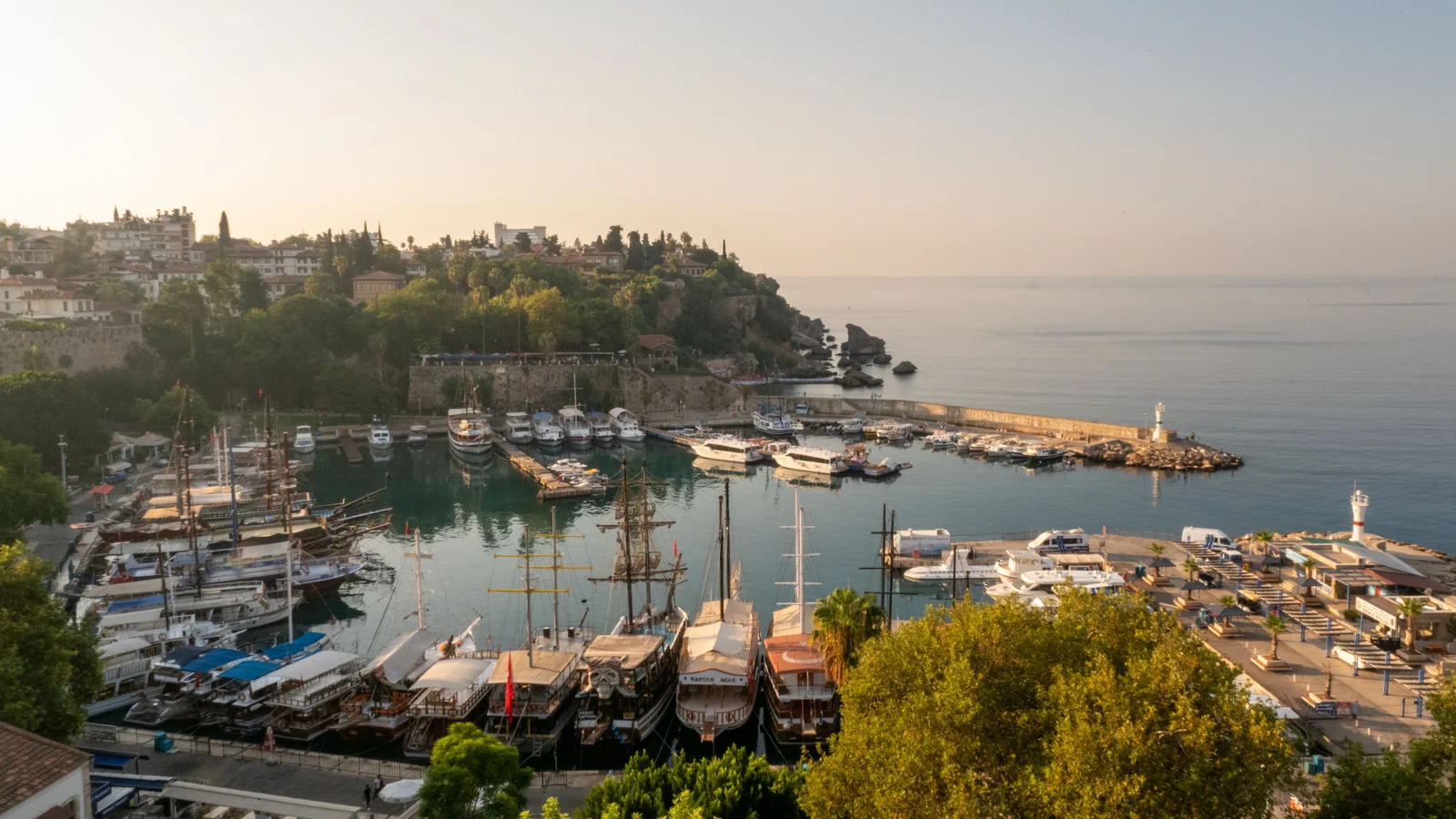 Boats in Antalya’s old harbour below Kaleiçi’s green headland in warm evening light.
