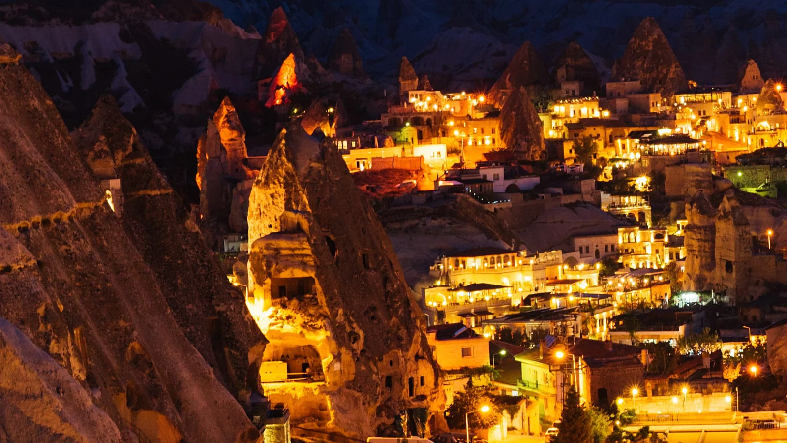 Illuminated rock-cut buildings and a tall fairy chimney among Göreme’s hillside houses at dusk.