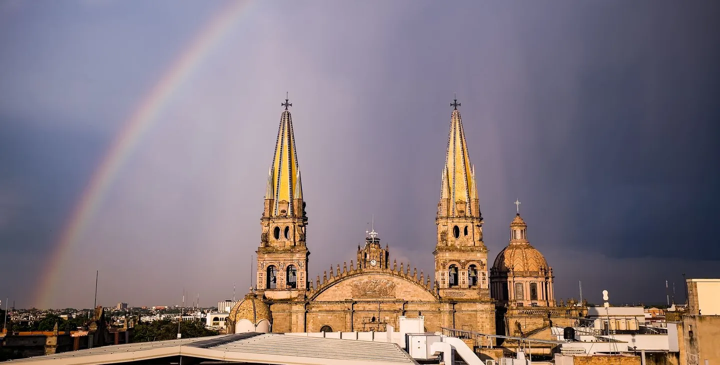 Twin yellow spires of Guadalajara Cathedral beneath a rainbow and storm clouds
