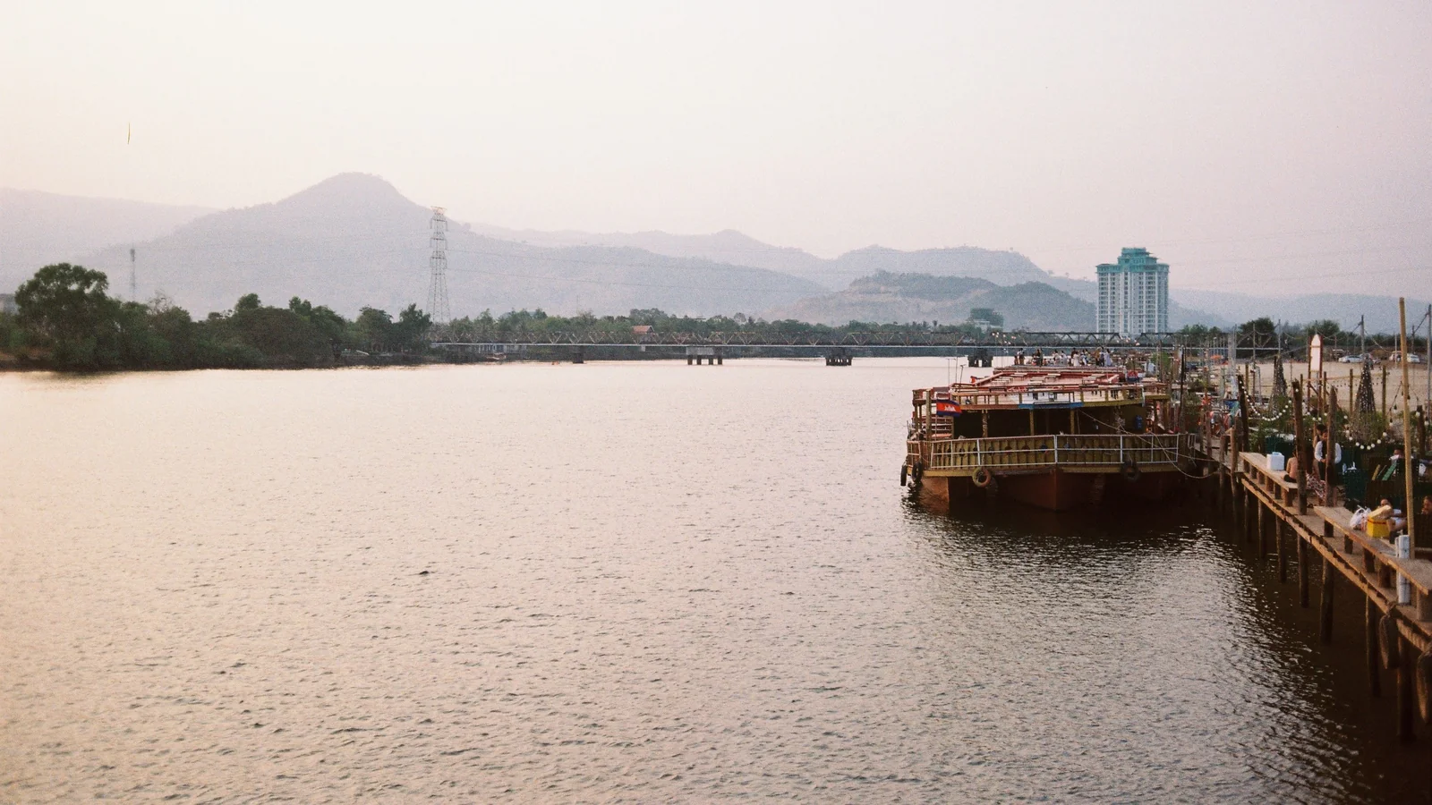 Boats moored beside Kampot’s riverfront, with the Old Bridge and mountains beyond.