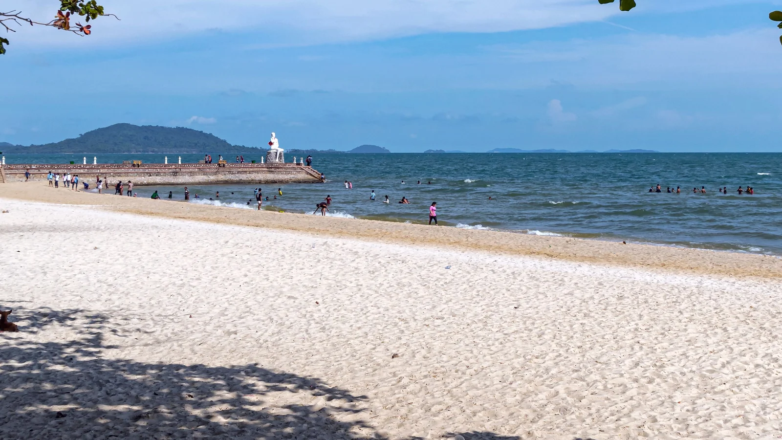 Kep Beach beneath leafy branches, with the White Lady statue on the seawall and islands offshore.