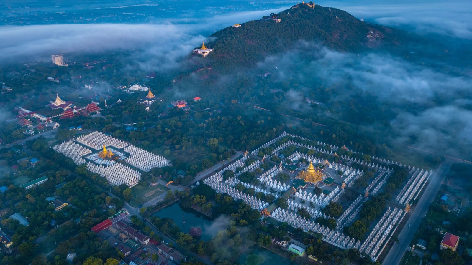 Mandalay Hill rising above the pagoda compounds of northeastern Mandalay, with Sandamuni’s compact white shrines in the foreground.