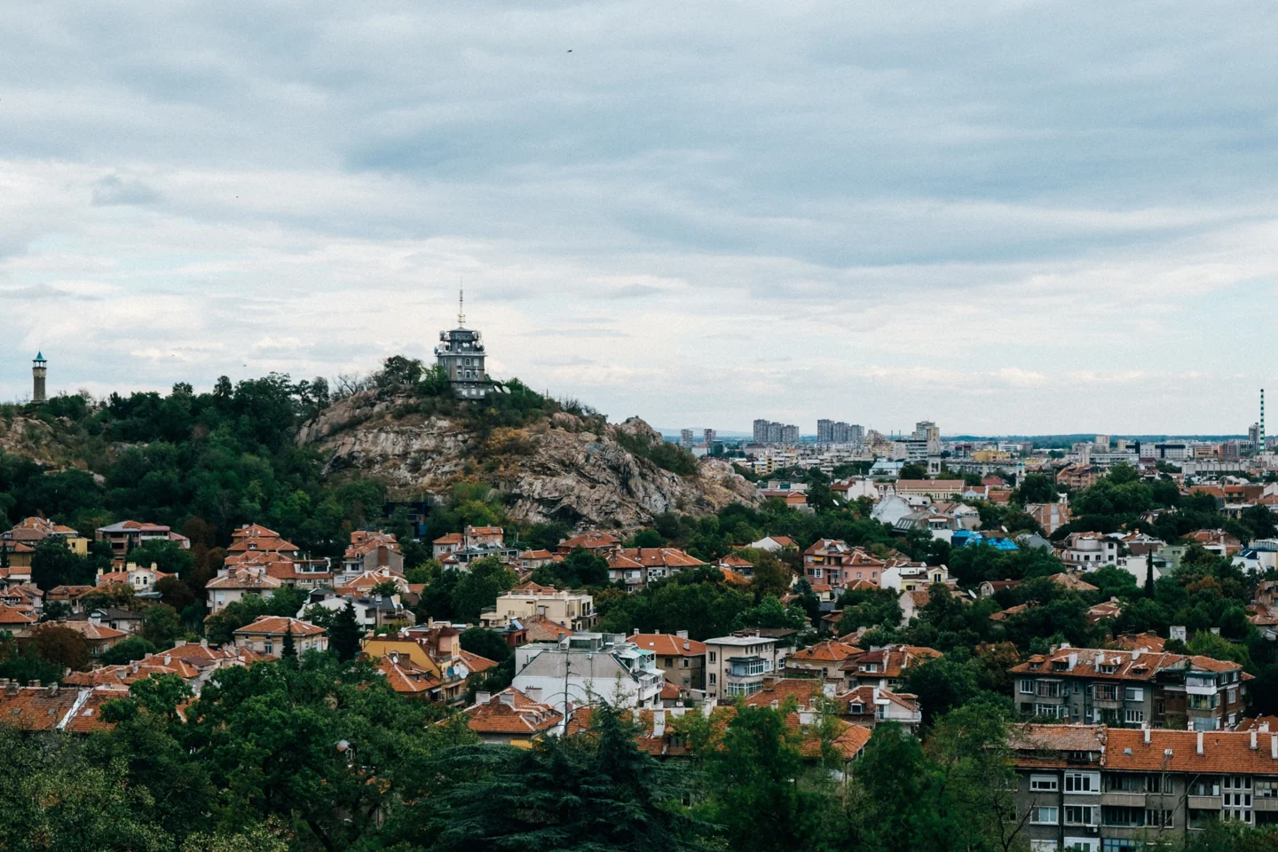 Plovdiv rooftops, green slopes and a hilltop monument under a cloudy sky